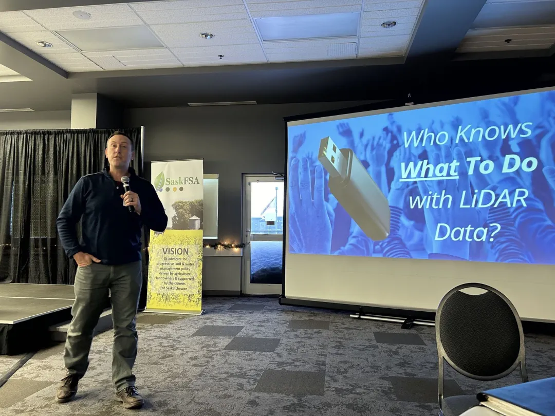 Middle-age man standing and holding a mic during a presentation; a SaskFSA promotional banner is in the background.